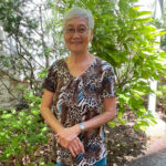 The image shows an elderly woman, Amy Wong, standing outdoors in a lush garden setting. She has short gray hair, glasses, and is smiling warmly at the camera. She is wearing a patterned blouse with earthy tones and light blue jeans. The garden around her is filled with greenery, including shrubs and small trees, creating a peaceful and serene atmosphere. Sunlight filters through the leaves, casting soft light on her and the surrounding plants, emphasizing the natural, calm environment.