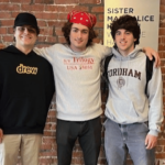 The image shows three young men standing side by side with their arms around each other, smiling at the camera. They are in front of a brick wall, creating a casual and relaxed atmosphere. The man on the left is wearing a black hoodie with the word "drew" on it and a black cap. The man in the middle is wearing a light gray hoodie with "s/v Trilogy" written on it and a red bandana. The man on the right is wearing a light gray hoodie with "Fordham" printed on it. They appear to be friends, sharing a lighthearted moment together. The overall vibe is friendly and casual.