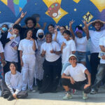 The image shows a group of young people standing in front of a colorful mural. They are all wearing white shirts, and many of them are making peace signs, posing happily for the photo. The mural behind them features vibrant artwork, including musical notes, abstract shapes, and other artistic designs, suggesting a creative or community-focused project. The group appears diverse, with a mix of men and women, some wearing hats or headscarves. The scene conveys a sense of teamwork, positivity, and involvement in a collaborative effort, related to the Summer Youth Employment Program.