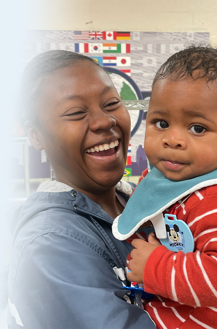 The image shows a smiling woman holding a baby boy in her arms. The woman is wearing a blue jacket and laughing joyfully, while the baby, dressed in a red and white striped outfit with a blue bib featuring Mickey Mouse, looks curiously at the camera. In the background, there is a wall decorated with various country flags and a large circular emblem. The image captures a moment of warmth and happiness, showcasing a close bond between the woman and the baby.