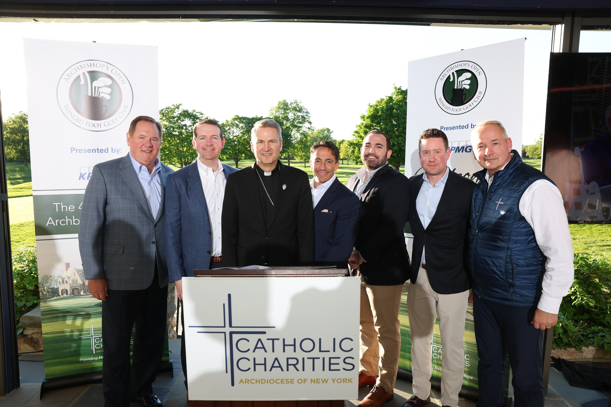 Seven people standing in behind the podium. In the middle is Archbishop Ronald Hicks in a black outfit with priest's collar. On the far-right is CEO Antonio Fernandez, with white shirt and blue vest. To the left of Archbishop is Sean Egan of KPMG. Behind them is a golf course, and two stand-up banners with a logo of a set of golf clubs and text that reads "40th Annual Archbishop's Open." Sign on the podium has a cross log and reads "Catholic Charities of the Archdiociese of New York"